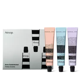 Un trio de baumes pour les mains Aesop, couleur peche, bleu pale et lilas, reposent sur leurs capuchons noirs, a cote d’une boite illustree gris argente.