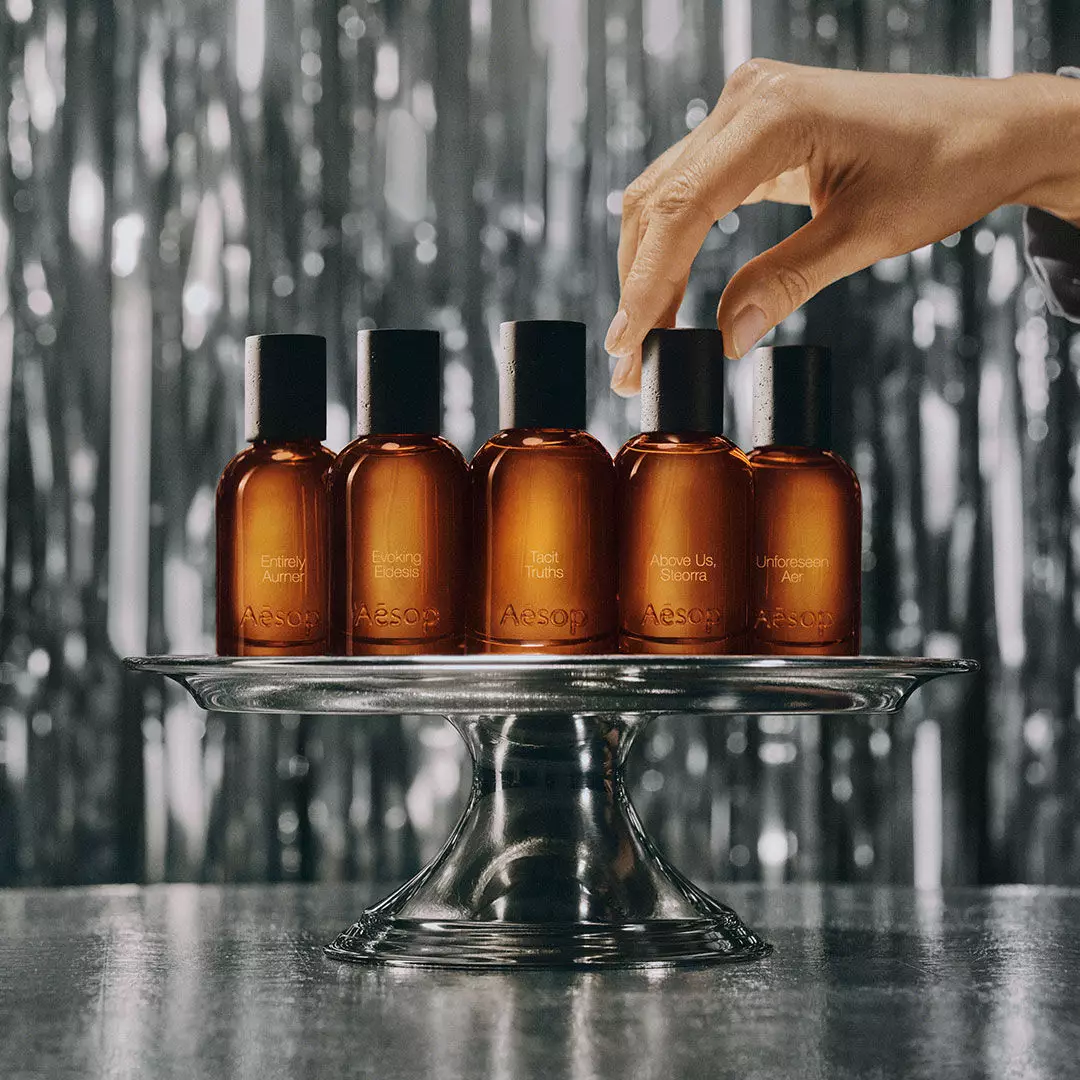 Deux boites Aesop, six petits flacons ambres et un objet blanc en forme de nez reposent sur un rebord reflechissant, surplombant des montagnes enneigees.