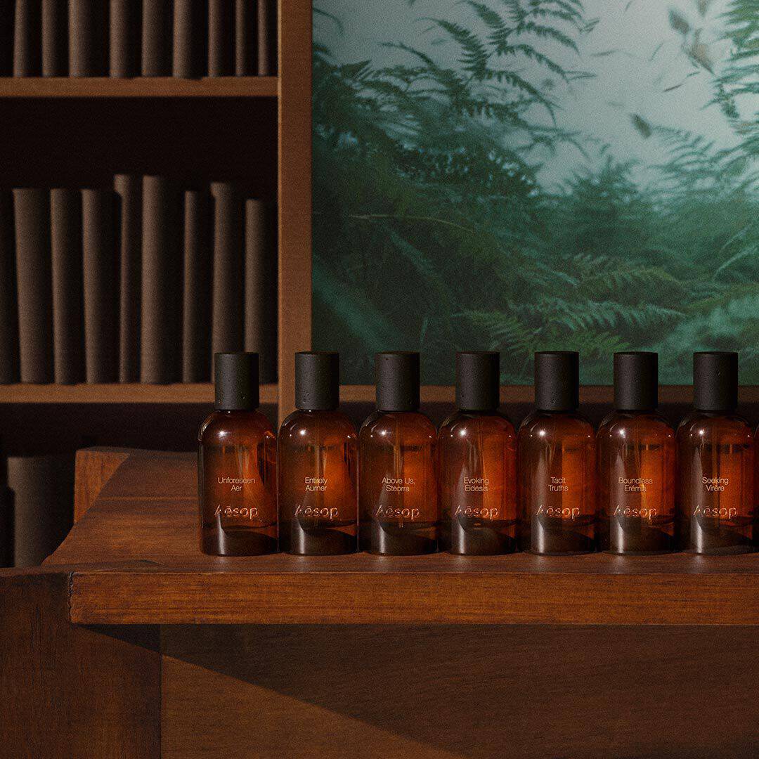 Rangee de flacons de fragrance Aesop ambres sur une table en bois, pres d’etageres et d’une image de fougere verte.