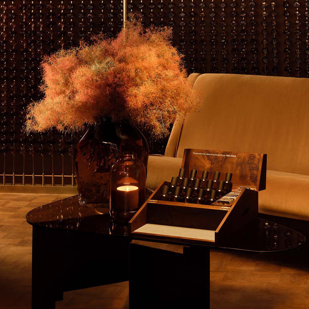 Nature morte d’interieur aux tons ambres, avec un canape, une table sombre, une composition florale sechee et des flacons de parfum presentes dans un coffret ouvert.