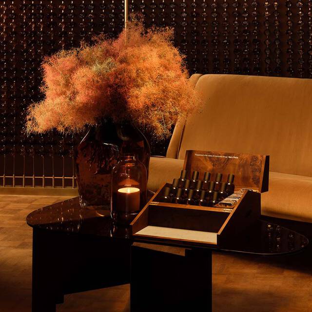 Nature morte d’interieur aux tons ambres, avec un canape, une table sombre, une composition florale sechee et des flacons de parfum presentes dans un coffret ouvert.