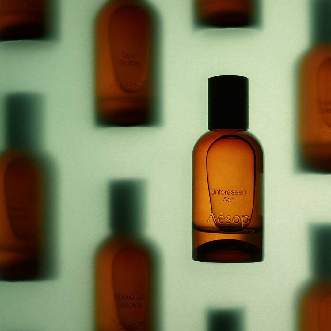 Flacon de fragrance Aesop Unforeseen Aer ambre, net au premier plan, devant des flacons ambres flous sur un fond vert pale.