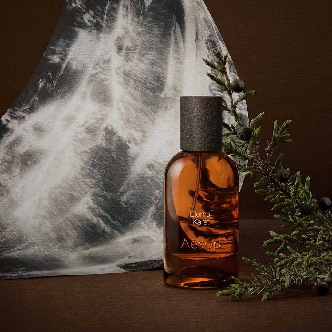Flacon de fragrance Aesop ambre, pose pres de brins de romarin et d’une image monochrome d’eau, sur une surface brun fonce.