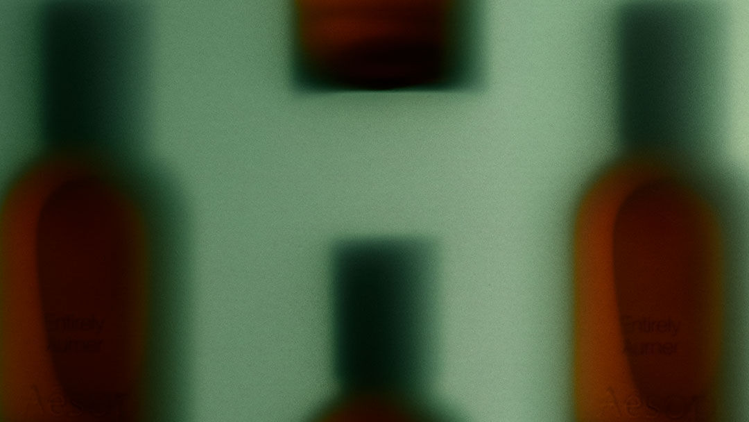 Flacons de fragrances ambrees aux contours flous, coiffes de bouchons sombres, sur fond vert pale, composant un motif horizontal repete et estompe.