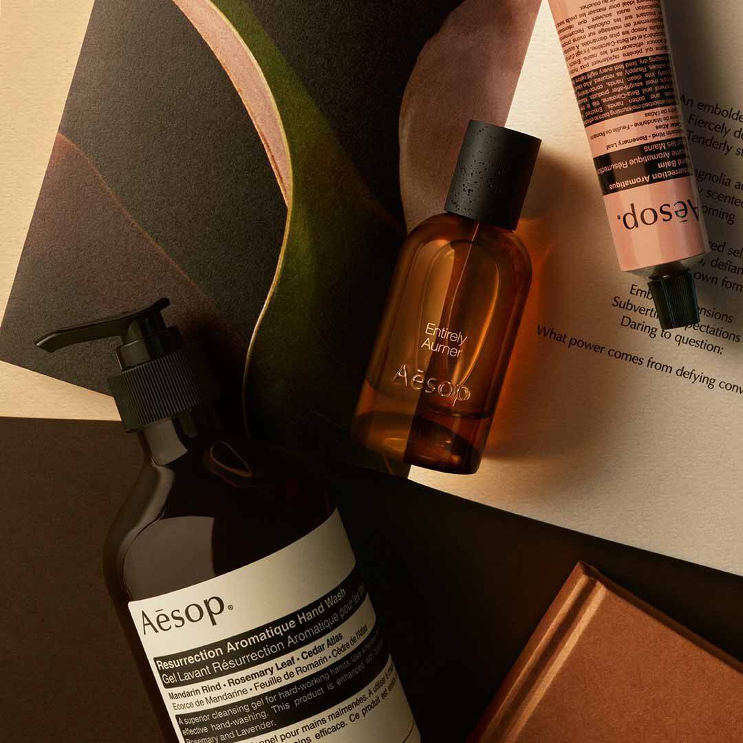 Flacon Aesop Entirely Aurner avec flacon-pompe et tube couleur peche, disposes sur des papiers et des formes sombres incurvees.