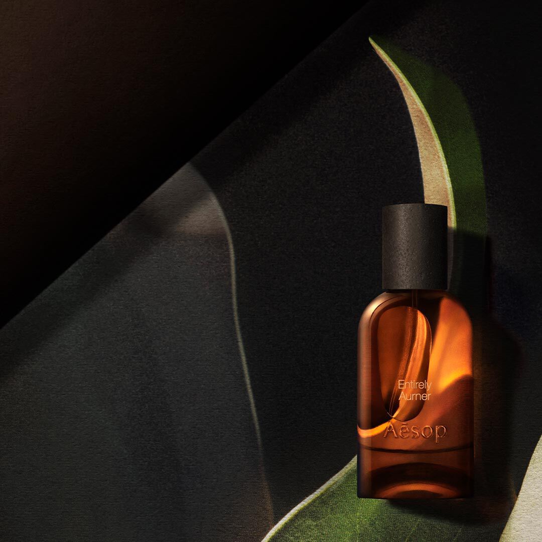 Flacon Aesop ambre solitaire, baigne d’une lueur chaleureuse, encadre de formes courbes evoquant des feuilles, dans des tons vert fonce, creme et noir.