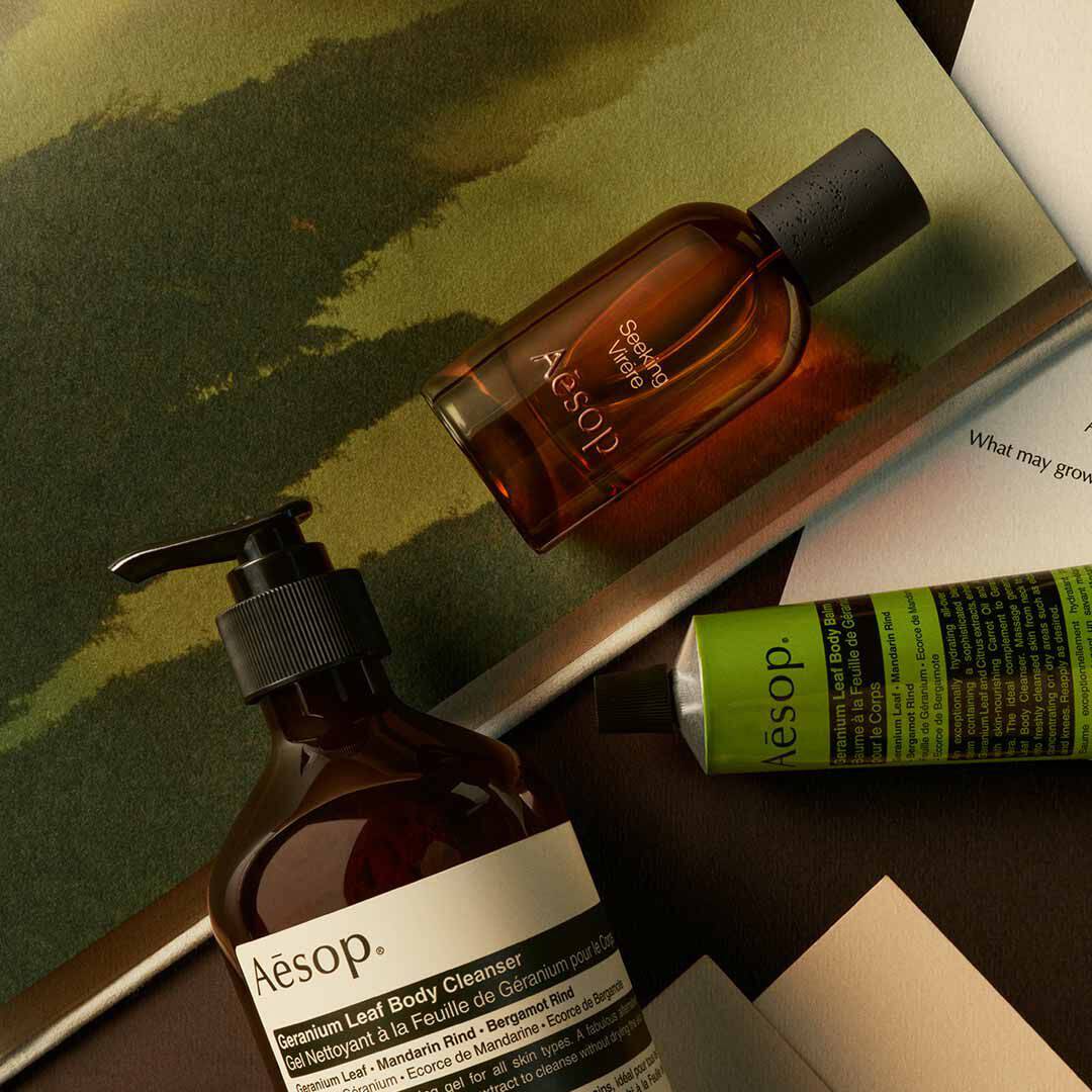 Flacon Aesop Seeking Virere avec flacon-pompe et tube vert, disposes sur des papiers avec des visuels peints dans des tons verts.