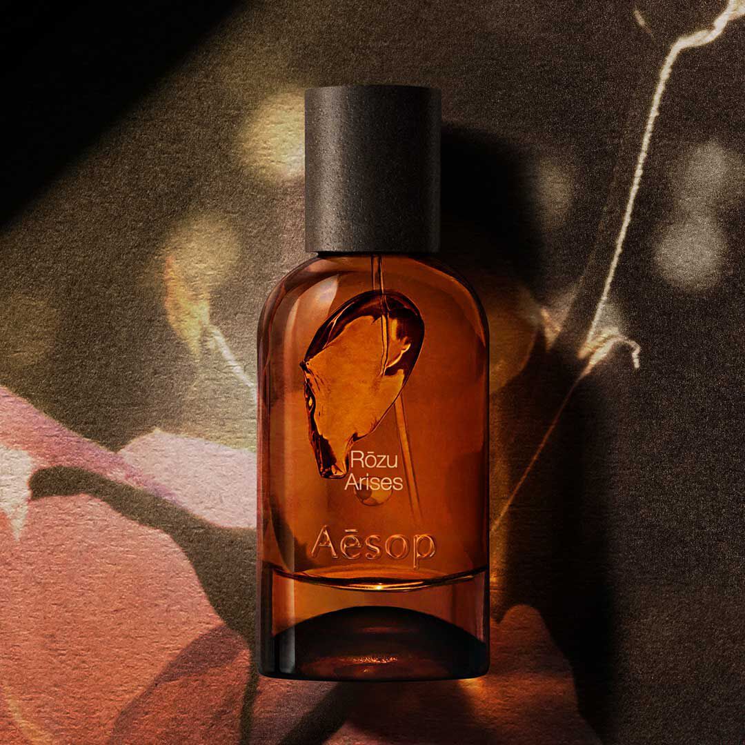 Flacon de fragrance Aesop Rozu Arises ambre sur une image florale sombre, baignee de lumiere chaude et d’ombre.