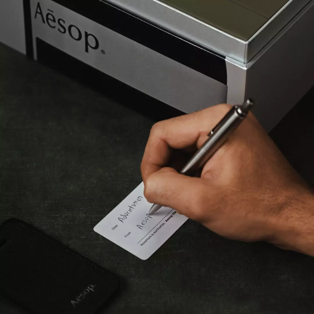 Une main ecrit sur une petite carte blanche, a cote d’une etiquette a bagage Aesop noire et d’un etui Aesop argente, sur un tissu vert fonce.