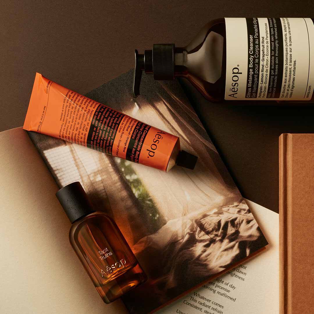 Flacon Aesop Tacit Truths avec flacon-pompe et tube orange, disposes sur des papiers avec une image monochrome.