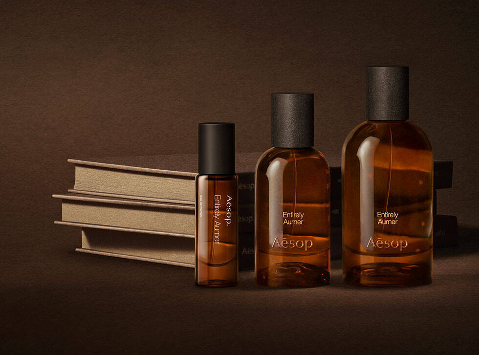 Trois flacons ambres de fragrances Aesop disposes a cote de livres creme empiles sur une surface brun fonce.