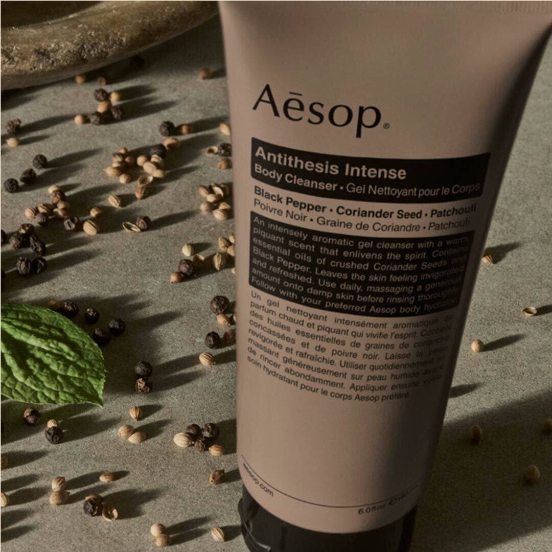 Aesop Antithesis Intense Body Cleanser tube beside black peppercorns, coriander seeds, and a green leaf.