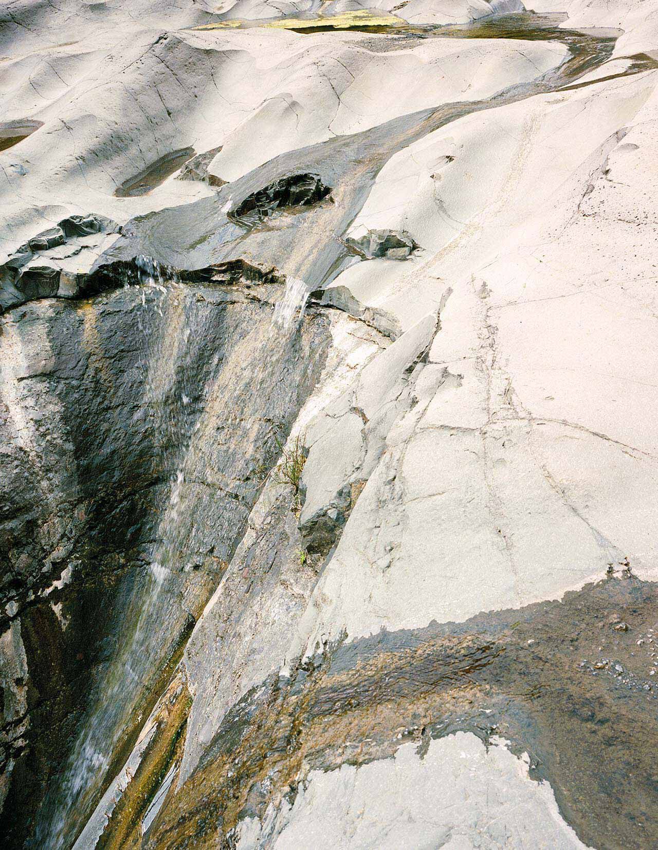 Surface minerale de pierre claire, marbree de stries blanches, grises et anthracite, aux textures superposees, traversee d’une fissure diagonale.