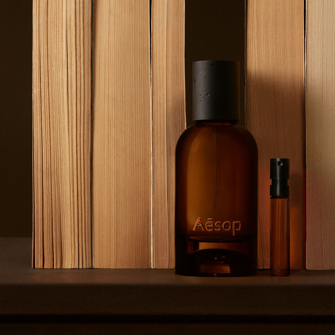 Flacon Aesop ambre et fine fiole disposes entre des planches verticales de bois clair, dans de profondes ombres brunes, selon une composition lineaire epuree.