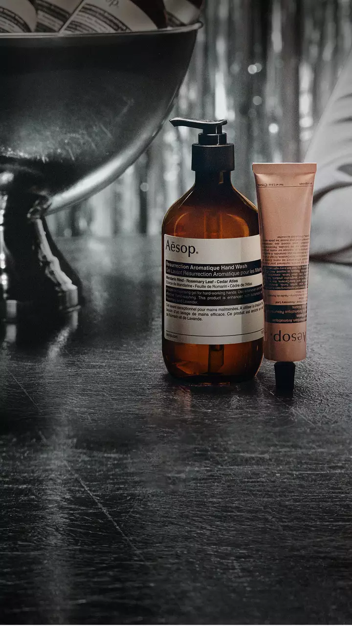 Un flacon-pompe Aesop ambre et un tube rose se tiennent a cote d’un grand bol sur pied, sur une table reflechissante, avec des serpentins argentes a l’arriere-plan.
