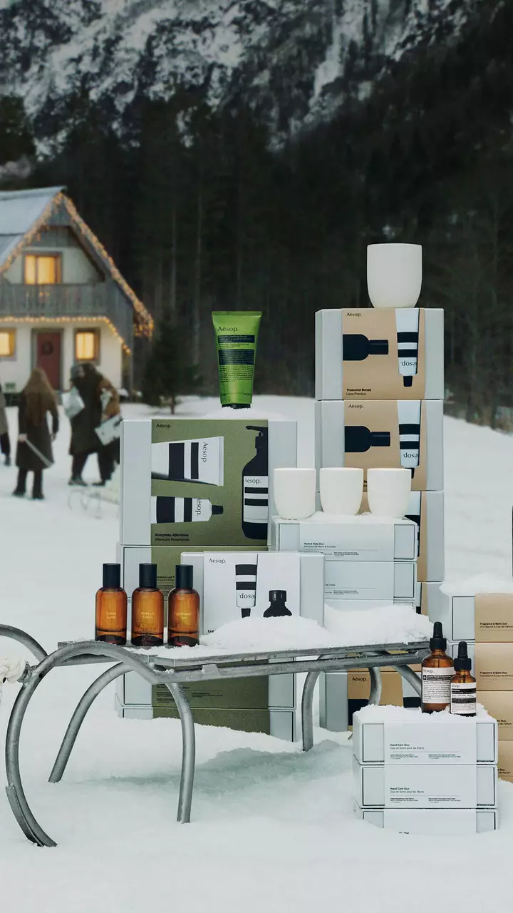 Des boites, flacons ambres, tubes colores et recipients blancs Aesop disposes autour d’une luge metallique dans la neige, avec des personnes a l’exterieur d’une maison eclairee a l’arriere-plan.