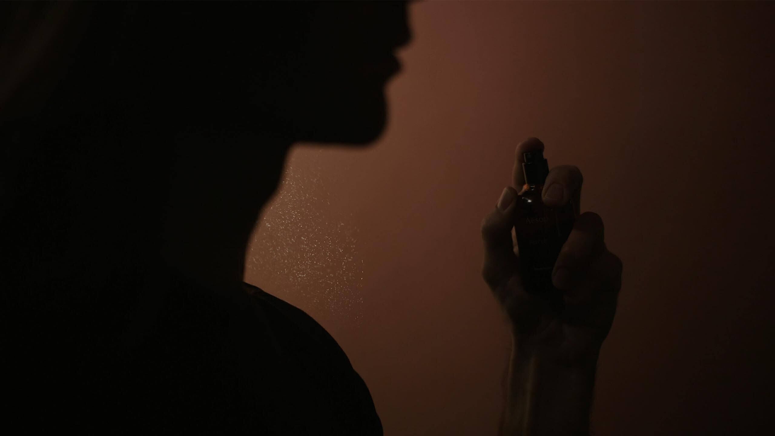 Silhouette d’une personne tenant un petit flacon de parfum pres de son visage, sur un fond ambre chaleureux.
