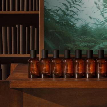 Rangee de flacons de parfum en verre ambre, coiffes de bouchons noirs, sur des etageres en bois a gradins, avec des livres et des fougeres en arriere-plan.