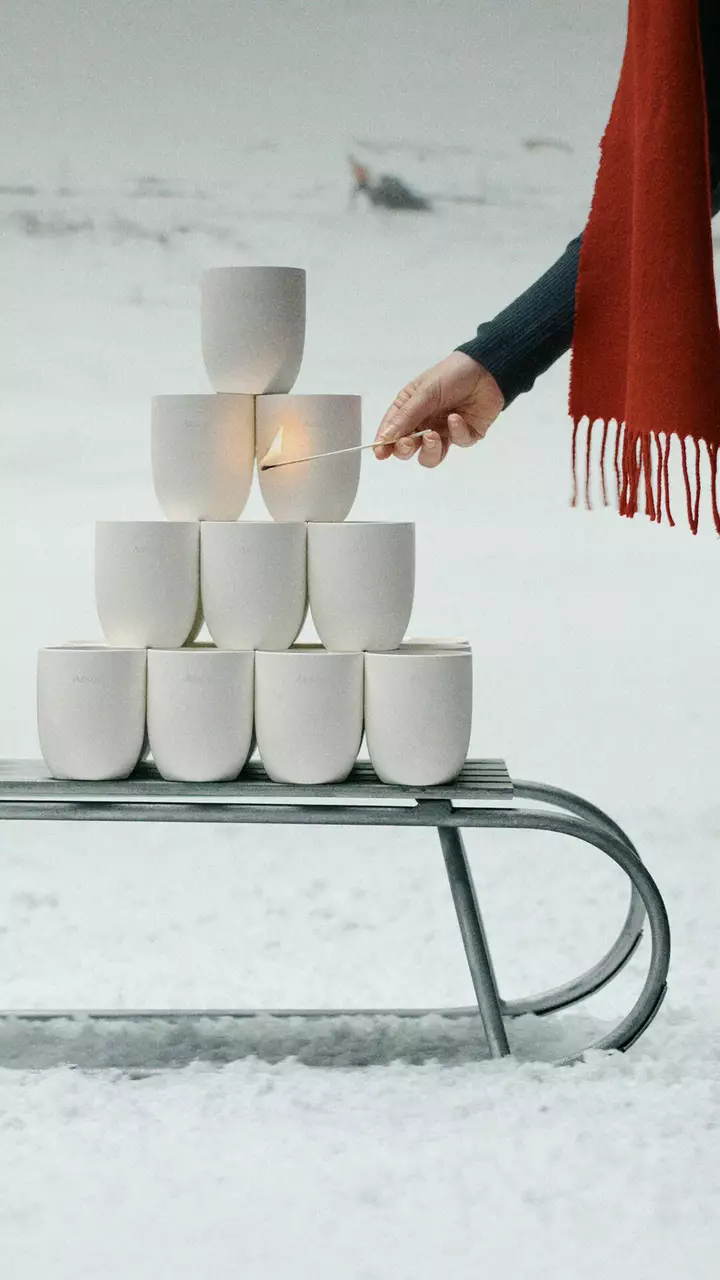 Une main tient une allumette enflammee pres de recipients blancs empiles sur un traineau metallique dans la neige, avec une echarpe rouge a franges sur la droite.